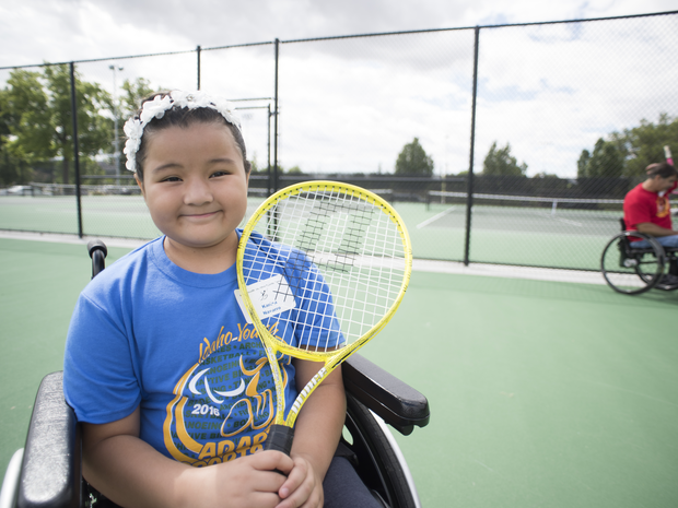 Young girl in wheelchair smiles while holding tennis racket.