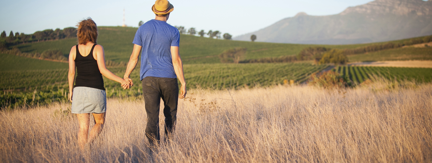 Two adults hold hands as they walk through a field of tall grass away from the camera