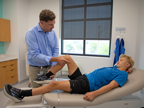 Orthopedic provider helps teenage boy bend knee on clinic bed.