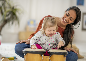 Woman holding toddler playing hand drum.