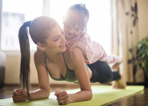 A mother planks on the floor, while her daughter lays on her back, smiling down