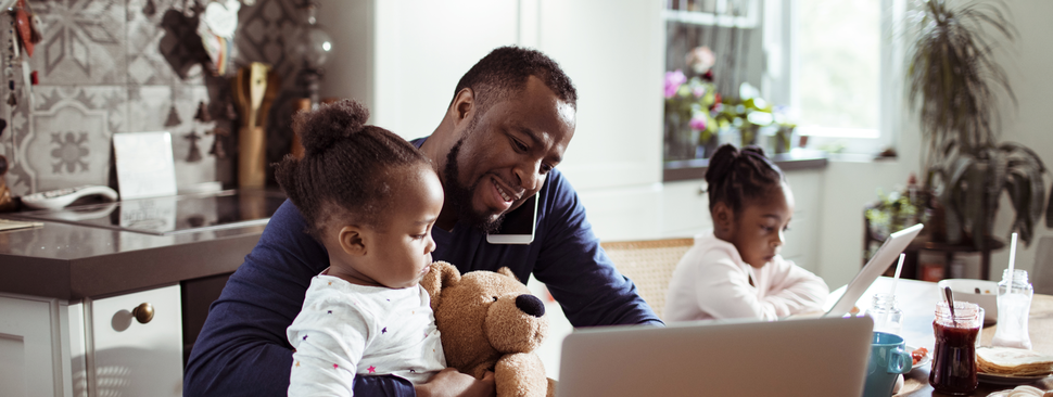 A dad works at the kitchen table while his 2 daughters sit with him