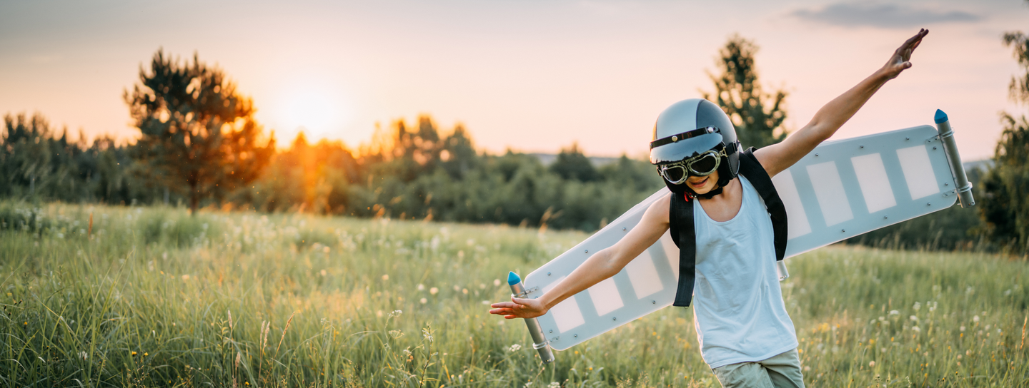 A boy runs through a field with his arms out while wearing model airplane wings