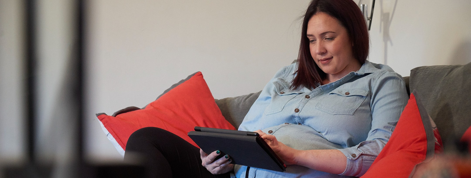 A pregnant woman sits on a couch, scrolling on a tablet