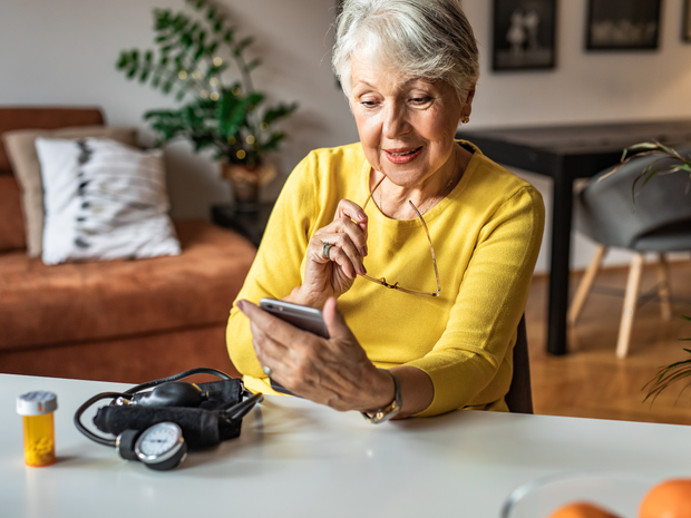 Older woman looks at phone with prescription bottle on kitchen table.