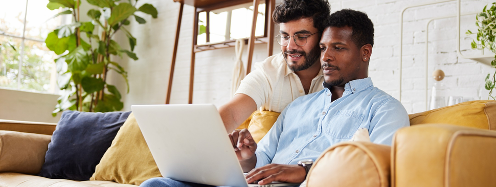 One man sits on his laptop on the couch, while another man leans over his shoulder