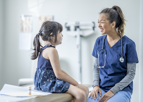 A young girl sits on an exam table while talking to a nurse