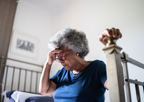 An older woman holds her head in pain while seated on her bed