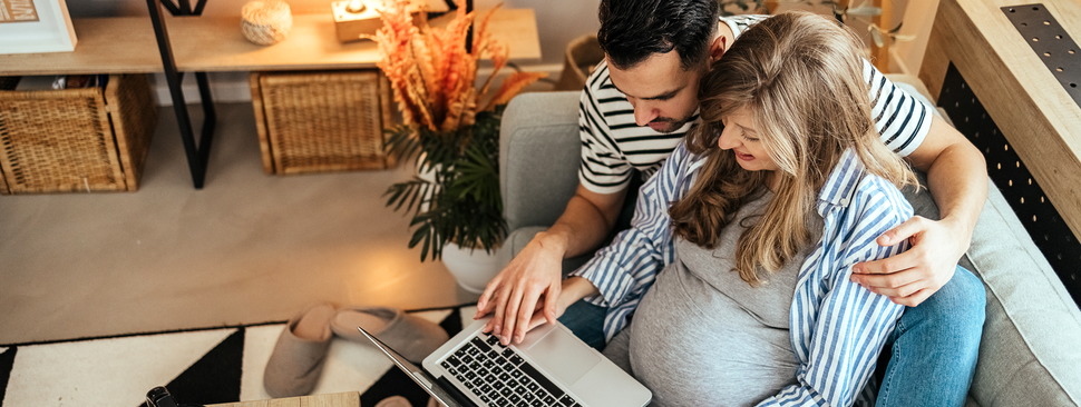 Pregnant couple sit together on the couch searching for a provider using a laptop