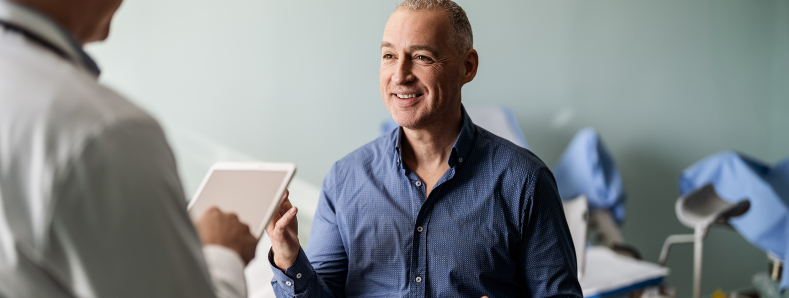 A bald man smiles as he talks to his doctor, who is holding a tablet