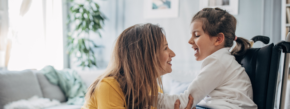 Mother and young daughter who is in wheelchair together in living room at home.