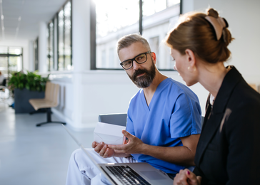 A woman dressed in a suit speaks with a doctor in scrubs while they sit in a hallway