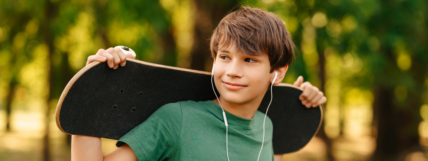 A boy wearing earbuds holding a skateboard.