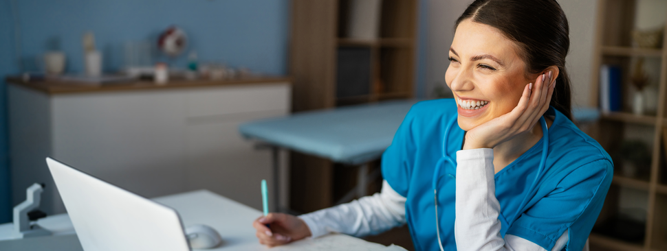 Cheerful female nurse in blue medical scrubs working on a laptop