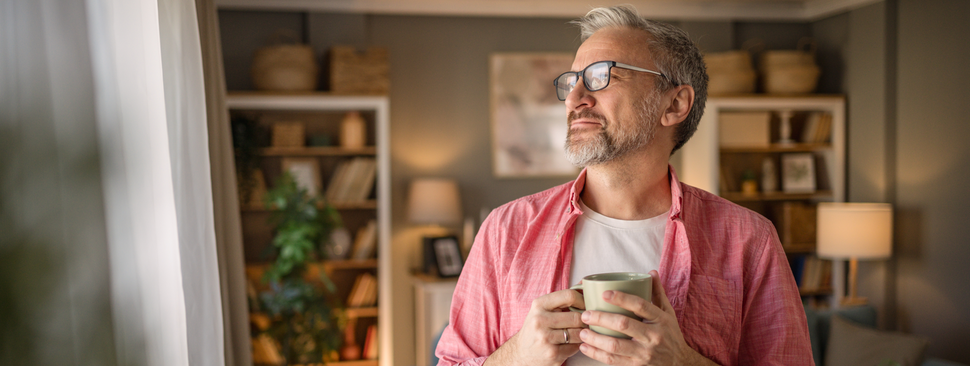 Mature man standing in the living room with a happy smile on his face and enjoying his morning coffee