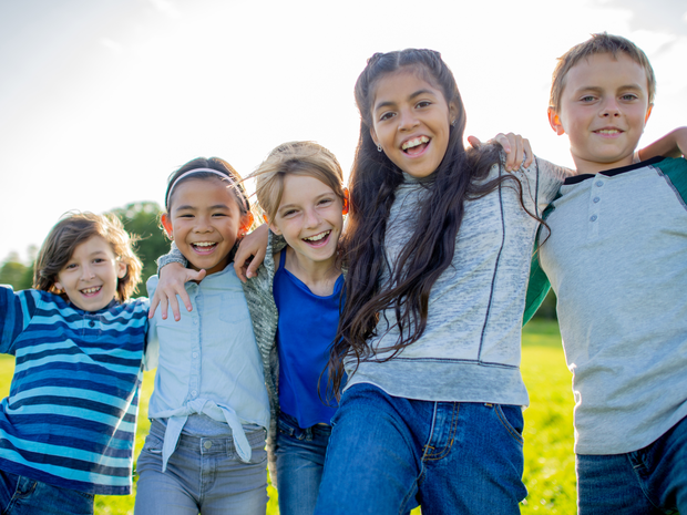 A small group of school-aged children stand side-by-side outside, with their arms around one another.