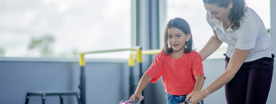 A young girl works on walking and keeping her balance with the help of her walker and her physical therapist.