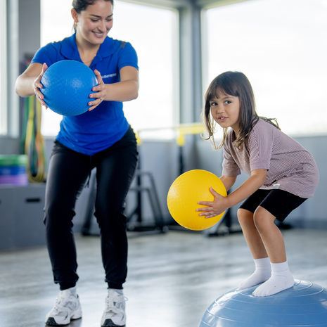 A physical therapist works with a young girl in a rehabilitation center, as they practice her balance and improve her core strength.