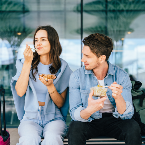 Two young adult friends, a Caucasian man and woman, eating snacks while sitting on the bench at airport terminal, outdoor.