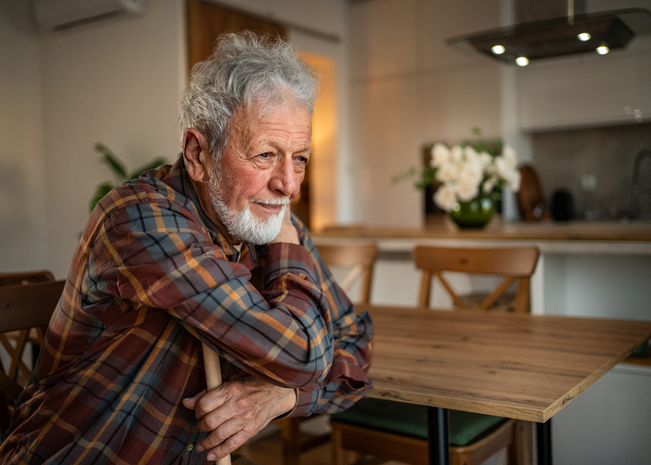 Elderly man sits at table with arms crossed.