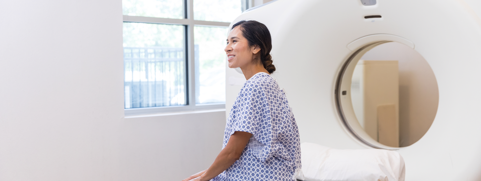 A smiling woman waits patiently for a medical exam near a CT scanner.