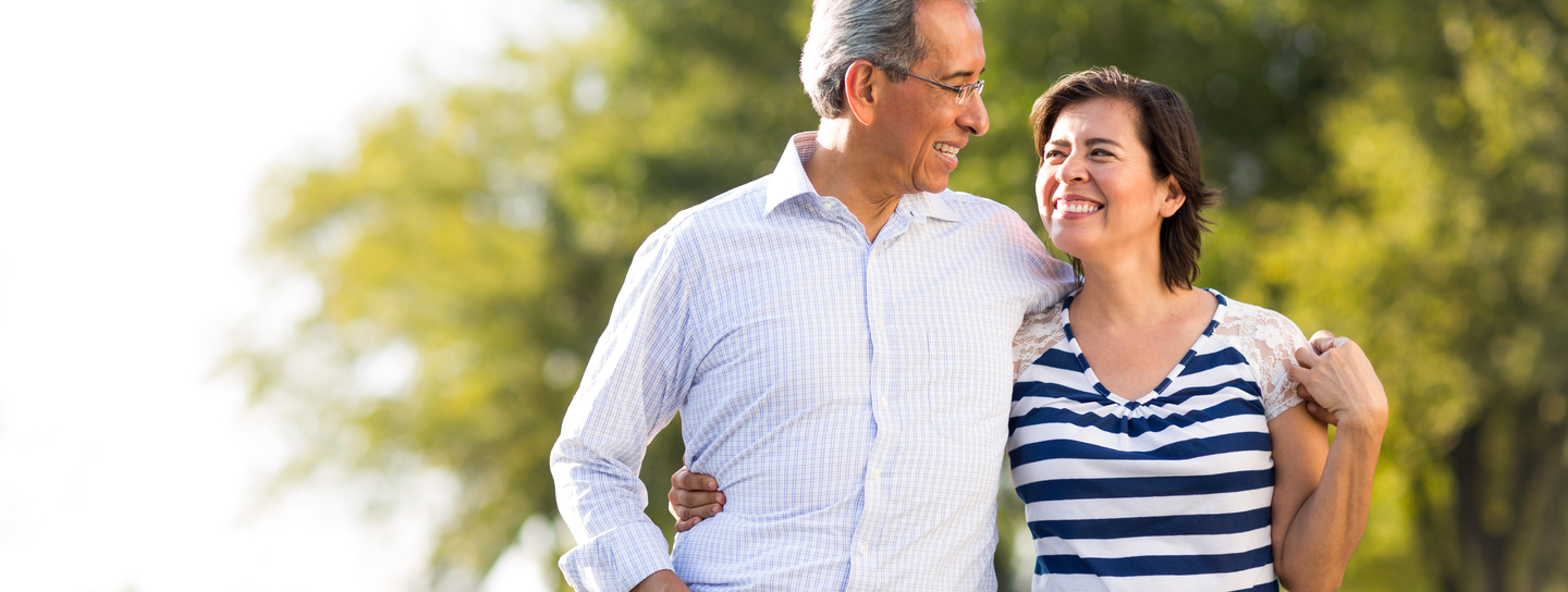 A mature couple walks outdoors, his arm is around her shoulders, as she holds onto his waist