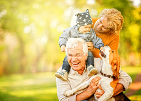 A grandfather smiles as his wife places their grandchild on his shoulders