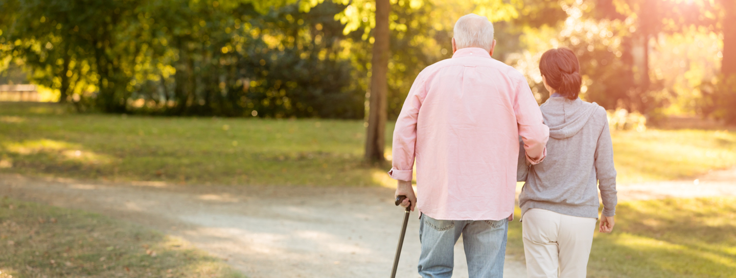 Older man uses a cane to walk outside with a young woman
