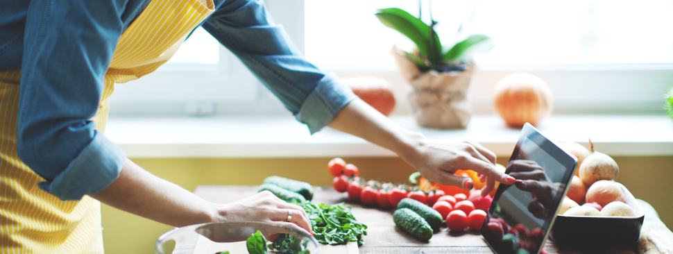 Woman in yellow apron looking up recipe on computer surrounded by vegetables 