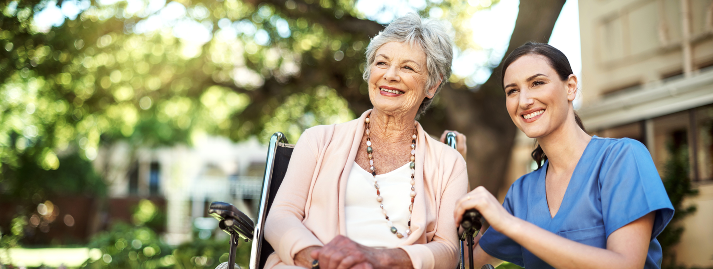 A mature woman in a wheelchair smiles next to a nurse in blue scrubs