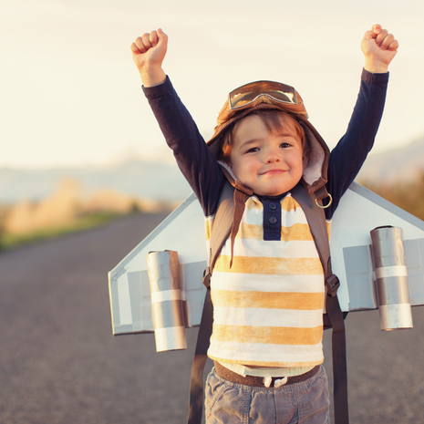 A young male child is standing on a rural road wearing a jet pack, flight cap and flying goggles. He wants to be a pilot when he grows up. He is smiling and is raising his arms. He loves aviation.