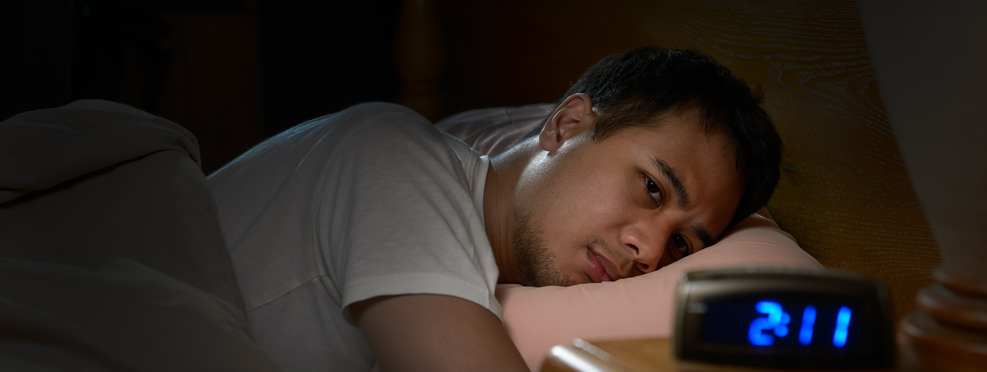 A man lays on his stomach in bed, looking at a digital clock that reads 2:11