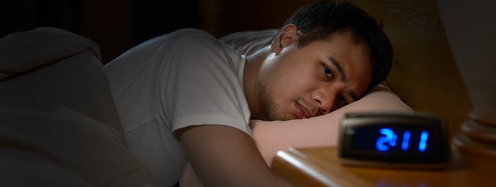 A man lays on his stomach in bed, looking at a digital clock that reads 2:11
