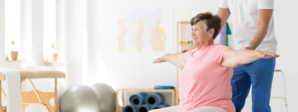 Older woman trying to maintain balance while sitting on a grey exercise ball, supported by a physical therapist