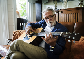 An older gentleman sits on his couch while playing guitar