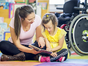 A young girl is sitting in front of her wheelchair. She is in an elementary school classroom with her helper. 