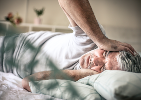 An older man lays on the bed, holding his head in pain