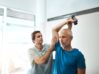 Middle-aged man works lifts small weights with provider support.
