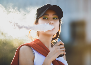 A woman holding a vape blows out a thick plume of smoke